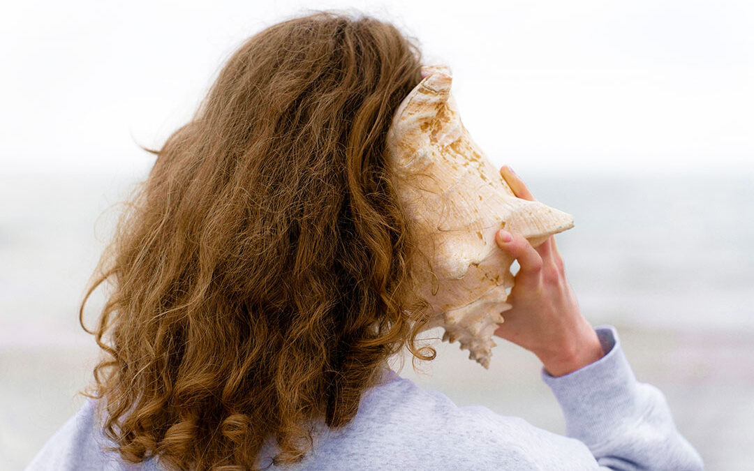 A woman seen from behind holding a conch to her ear.